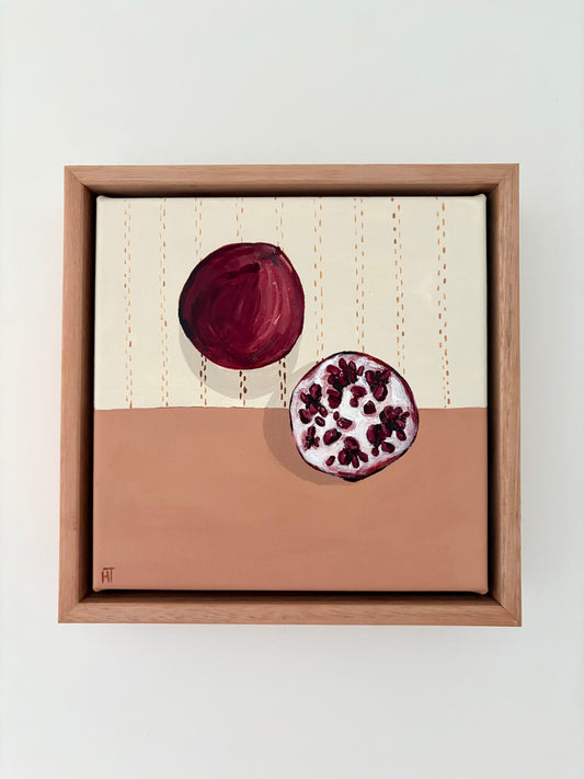Framed painting of a pomegrante on a halved cream and pink background. Cream section has brown stitching detail. 