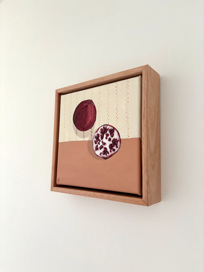 Framed painting of a pomegrante on a halved cream and pink background. Cream section has brown stitching detail. 