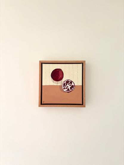 Framed painting of a pomegrante on a halved cream and pink background. Cream section has brown stitching detail. 