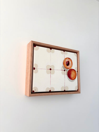Halved peach on a patterned table cloth in an oak frame. 