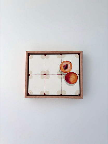 Halved peach on a patterned table cloth in an oak frame. 
