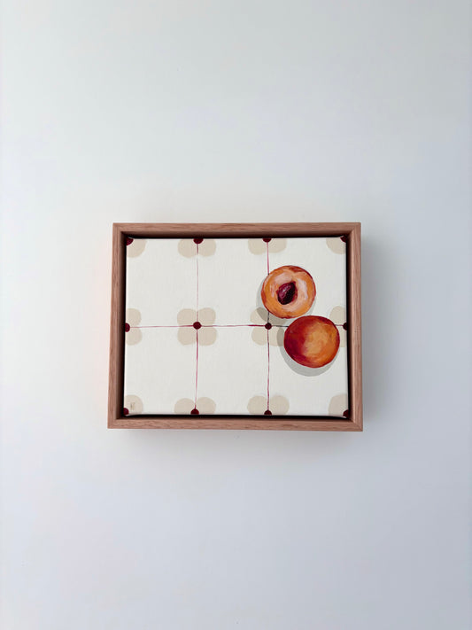 Halved peach on a patterned table cloth in an oak frame. 